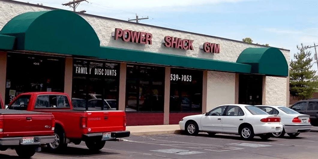 Storefront of Power Shack Gym, featuring red signage and green awnings. A red truck and white car are parked in front.