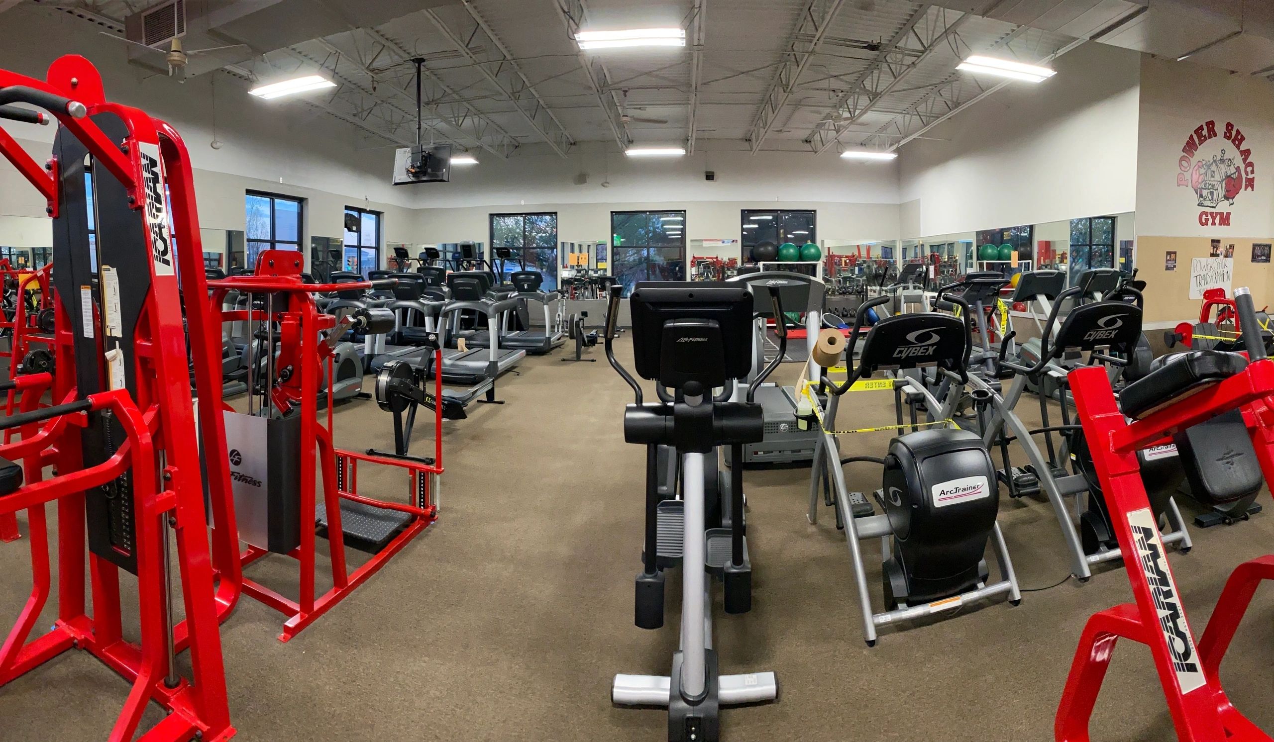 A gym filled with various cardio and weight machines, including treadmills, ellipticals, and red strength training equipment, under bright fluorescent lighting.