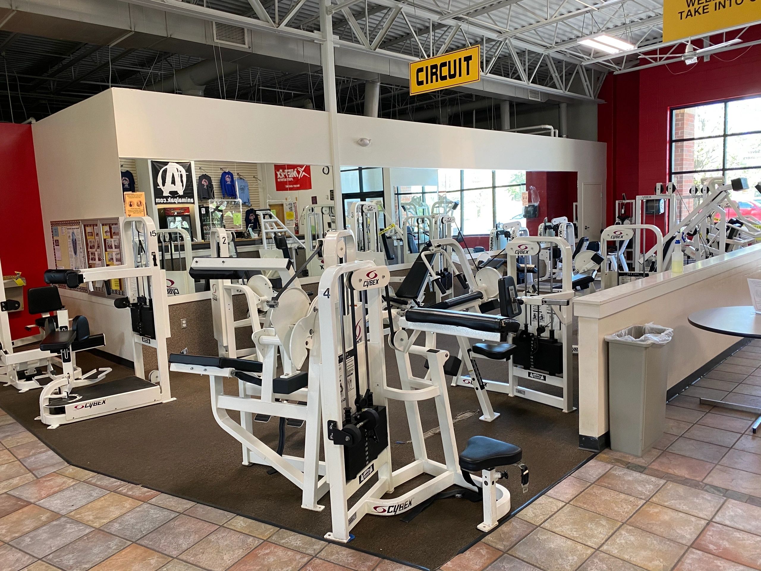 Gym interior with multiple weight machines arranged in a circuit layout. Walls display fitness posters, and there's a clothing rack. Ceiling has exposed beams and fluorescent lighting.