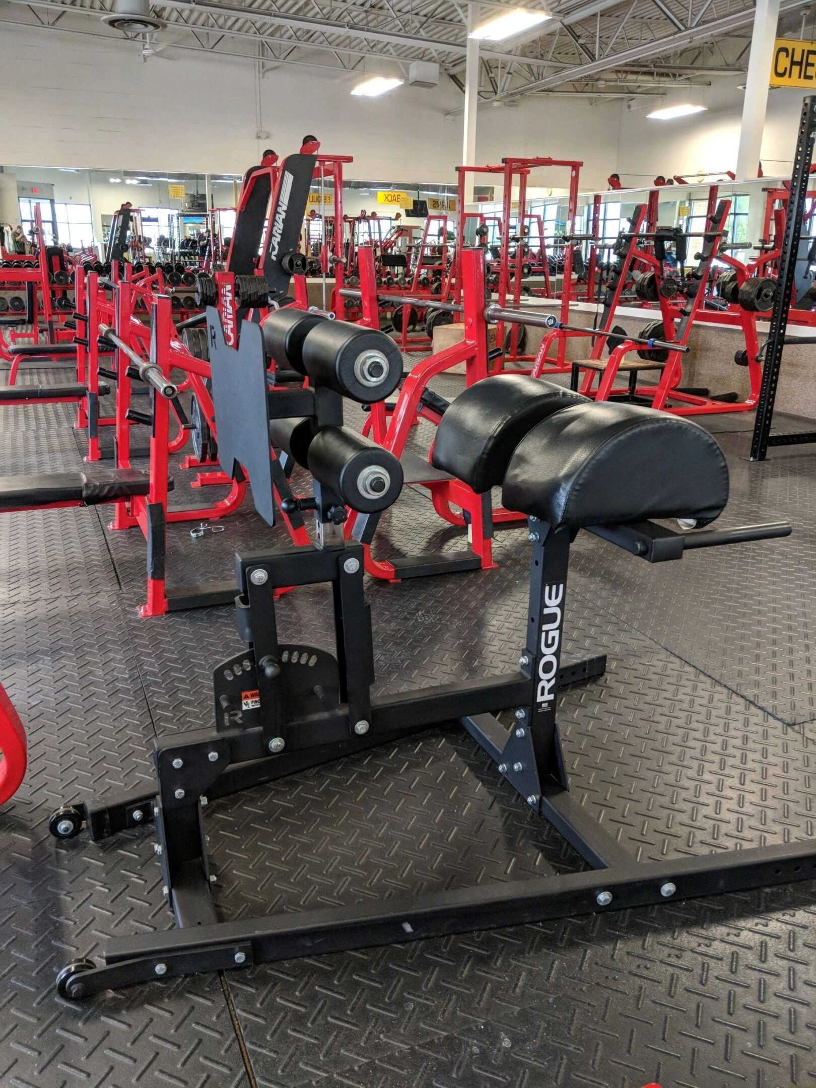 Gym equipment in a fitness center, featuring a black and red Rogue hyperextension bench on a checkered rubber floor, surrounded by red weightlifting racks and benches.