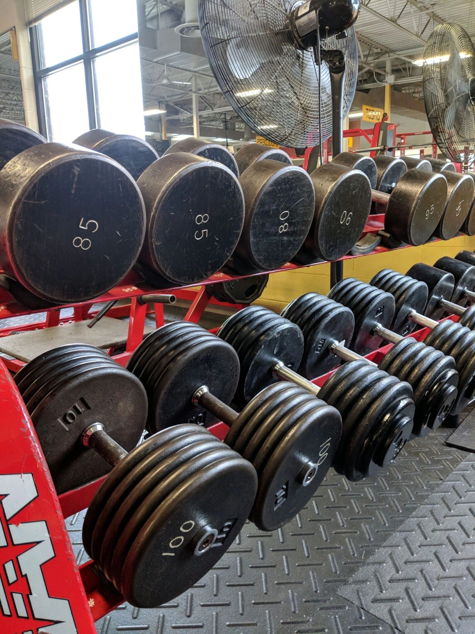 Dumbbells on a rack in a gym, ranging from 80 to 100 pounds, with large industrial fans visible in the background.