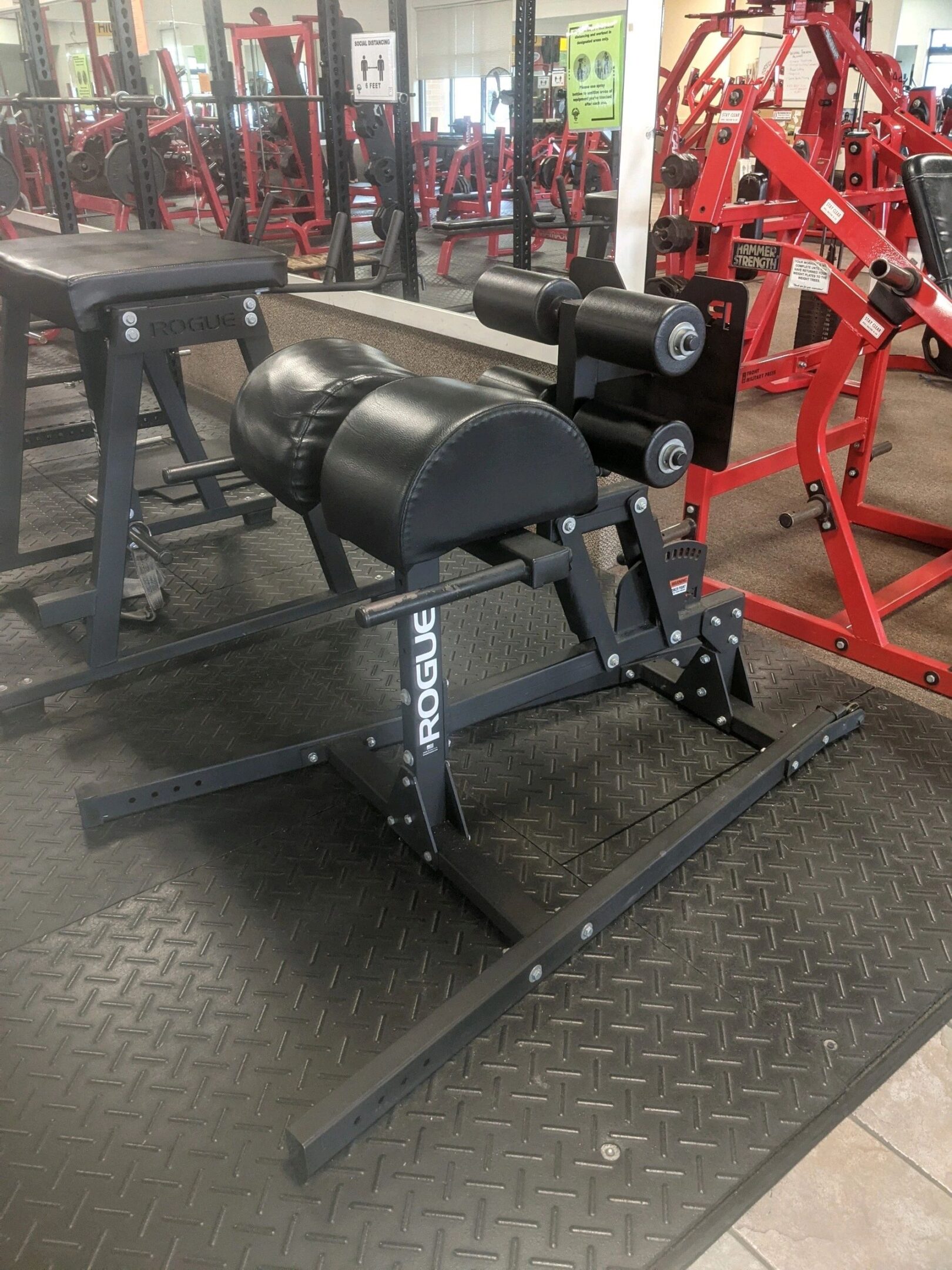 Gym equipment setup with a Rogue glute-ham developer on a rubber mat. Red and black exercise machines are visible in the background.