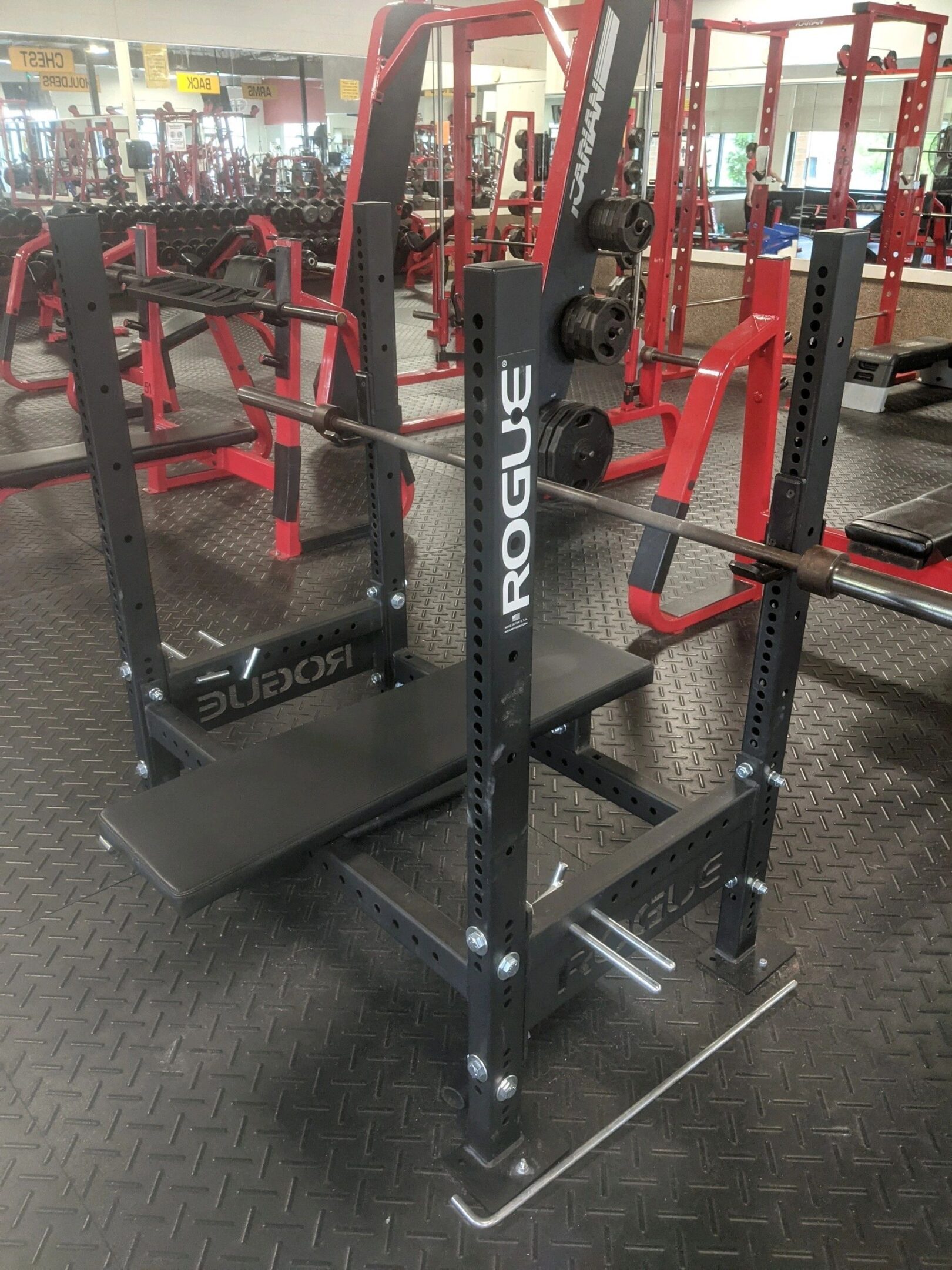 A gym bench press setup with a Rogue rack, barbell, and weight plates on a rubber floor.