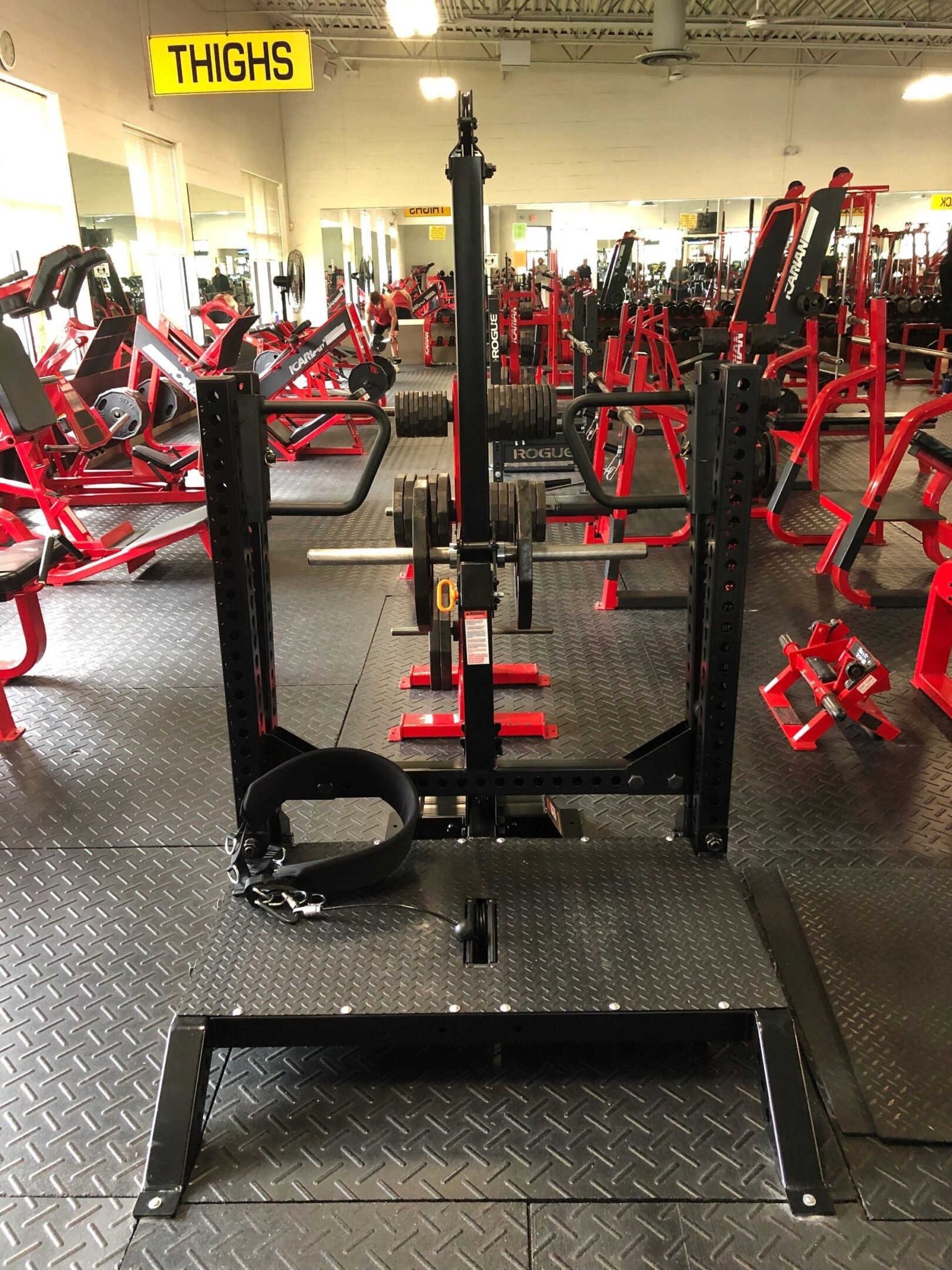 Smith machine in a gym, surrounded by other weightlifting equipment. A sign above reads "THIGHS.