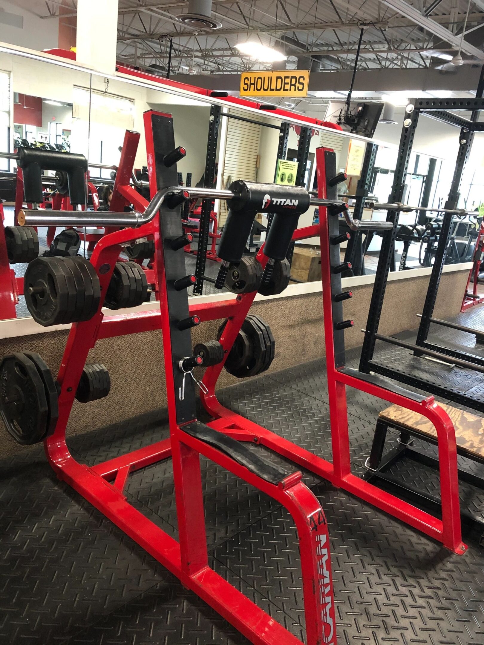 Gym equipment setup with a red squat rack, weights, and labeled "Shoulders" sign above. Mirrors and other racks in the background.