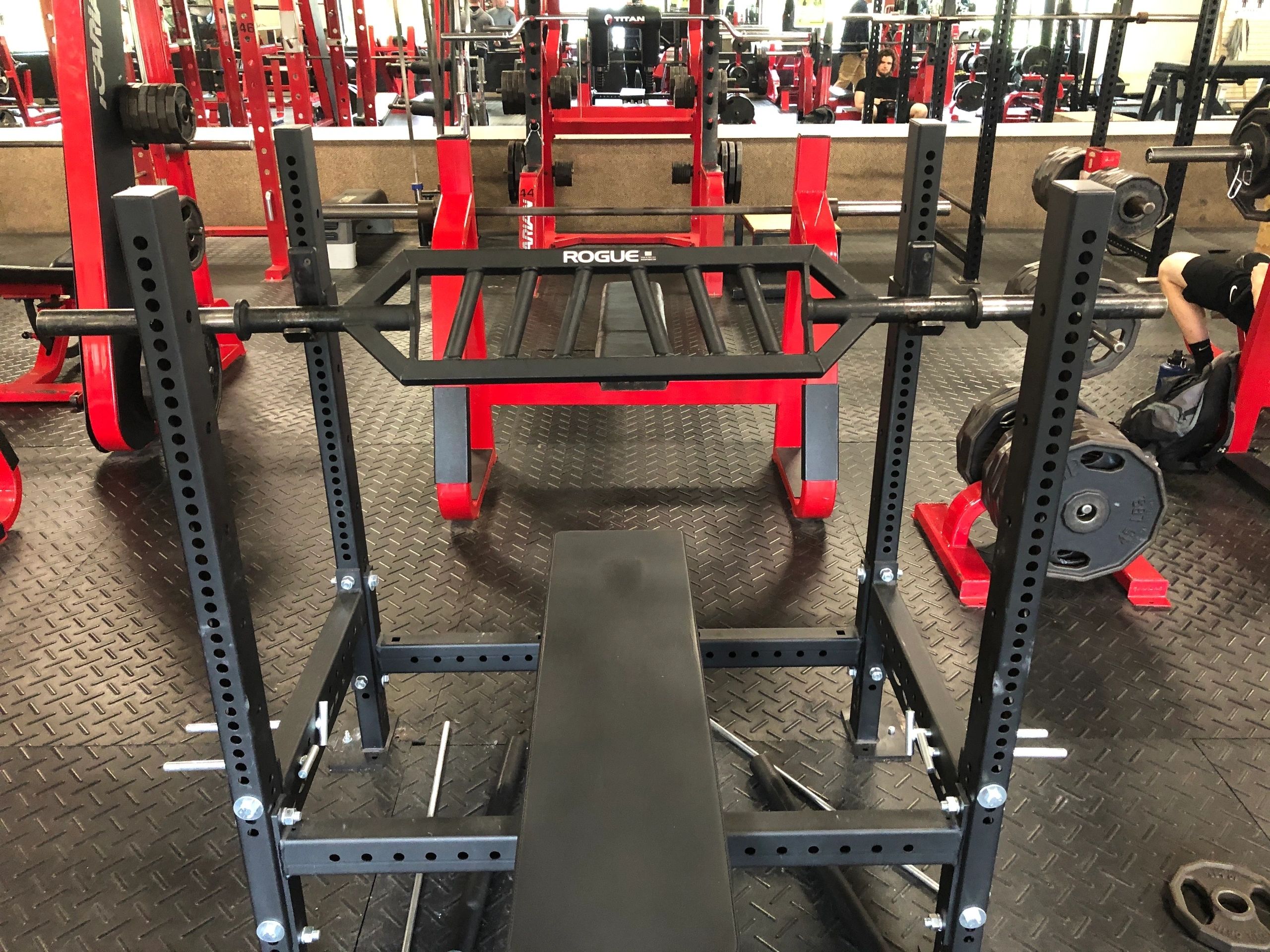 A gym setup featuring a squat rack with a barbell, weights, and a bench on a rubber mat floor. Red and black gym equipment is visible in the background.