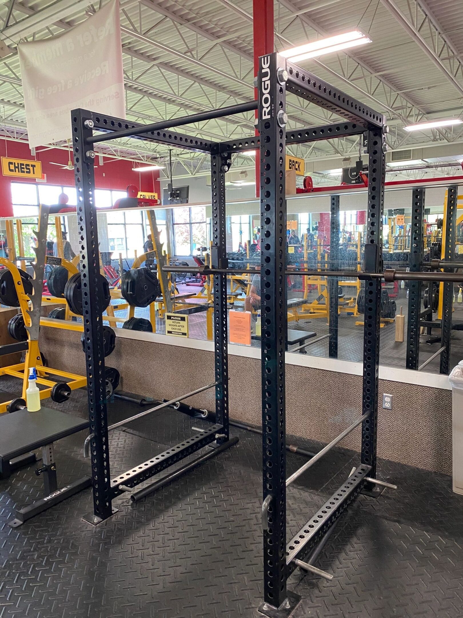 A black Rogue power rack in a gym, surrounded by weight benches, mirrors, and equipment.