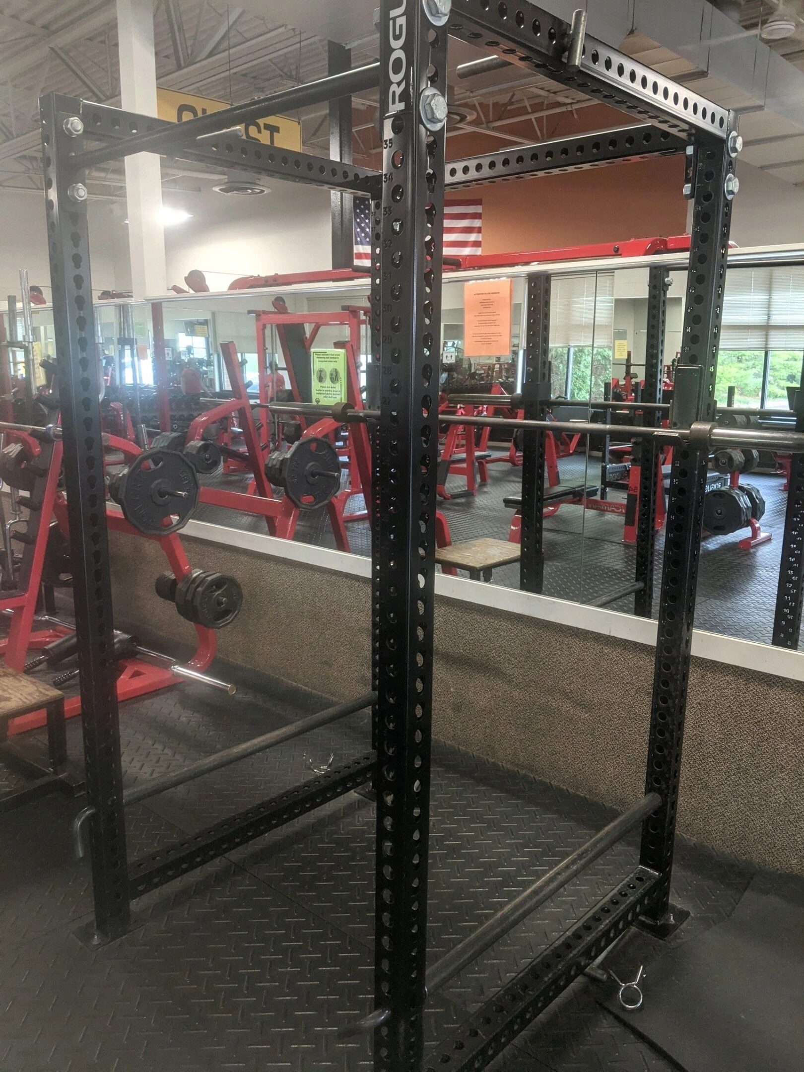 A black power rack with a barbell is set up in a gym. Reflective mirrors and other gym equipment are visible in the background.
