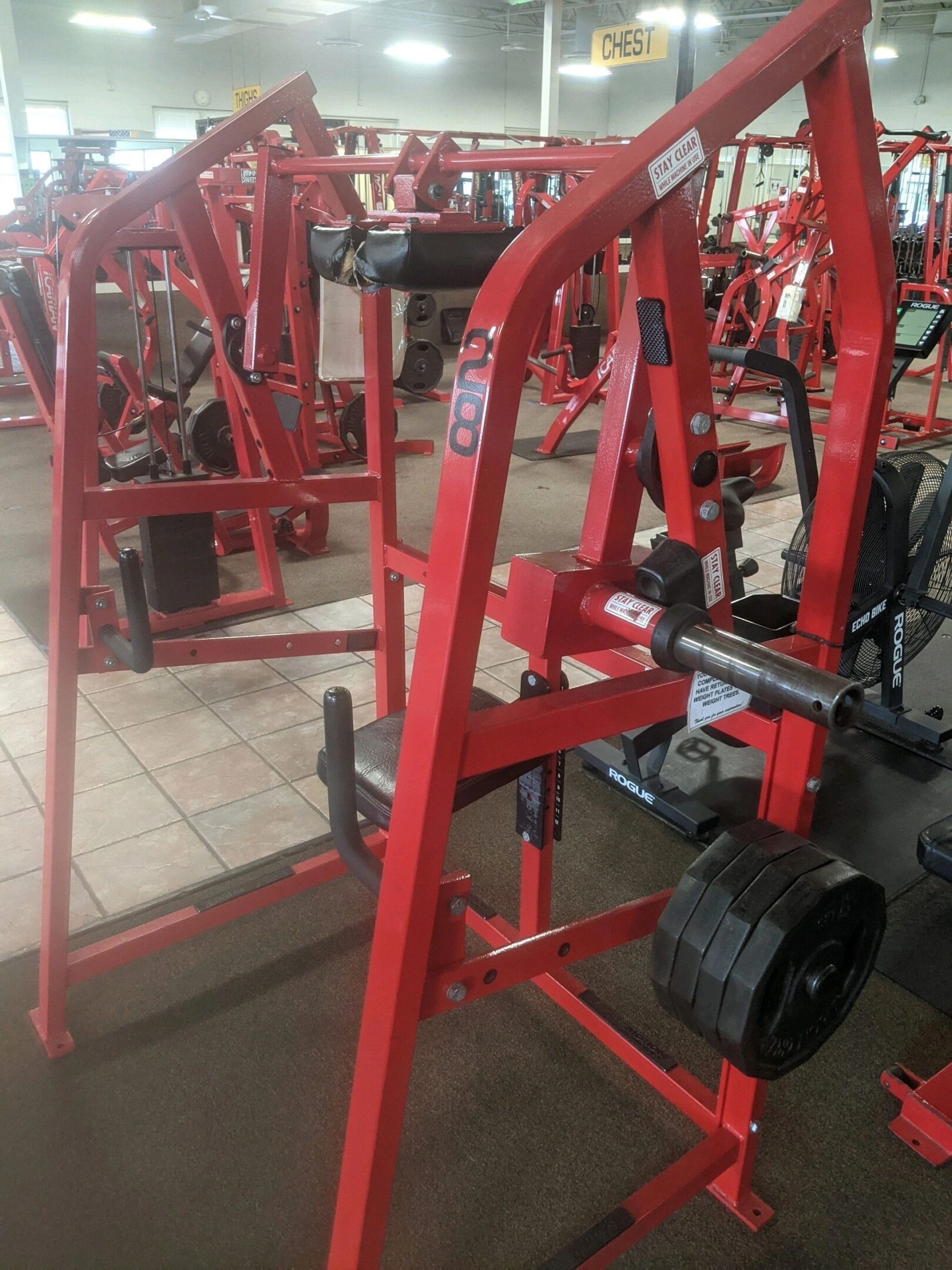 Red gym equipment, likely a chest press machine, with weights loaded on the side, placed in a fitness center.