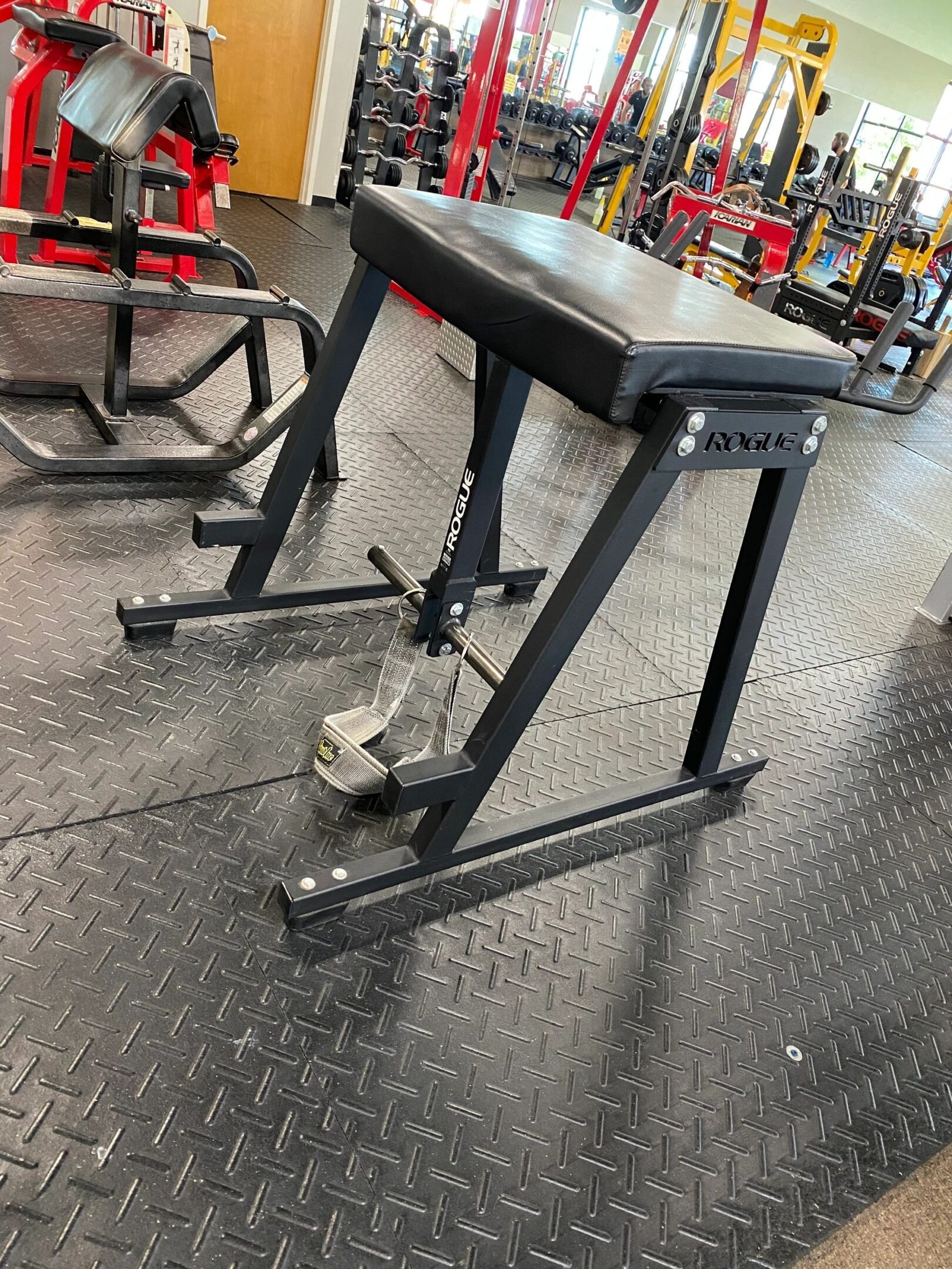 Gym equipment on a rubber floor, featuring a Rogue hyperextension bench with black padding and metal frame.