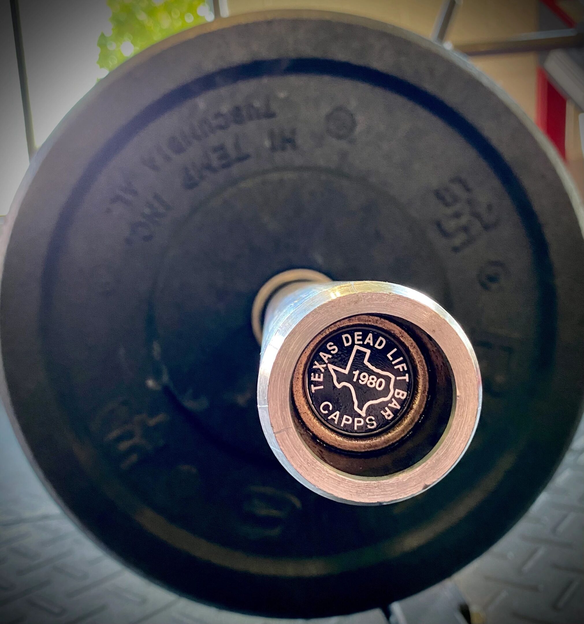 Close-up of a Texas deadlift bar with the label "Capps 1980" on the end cap, resting on a gym floor.