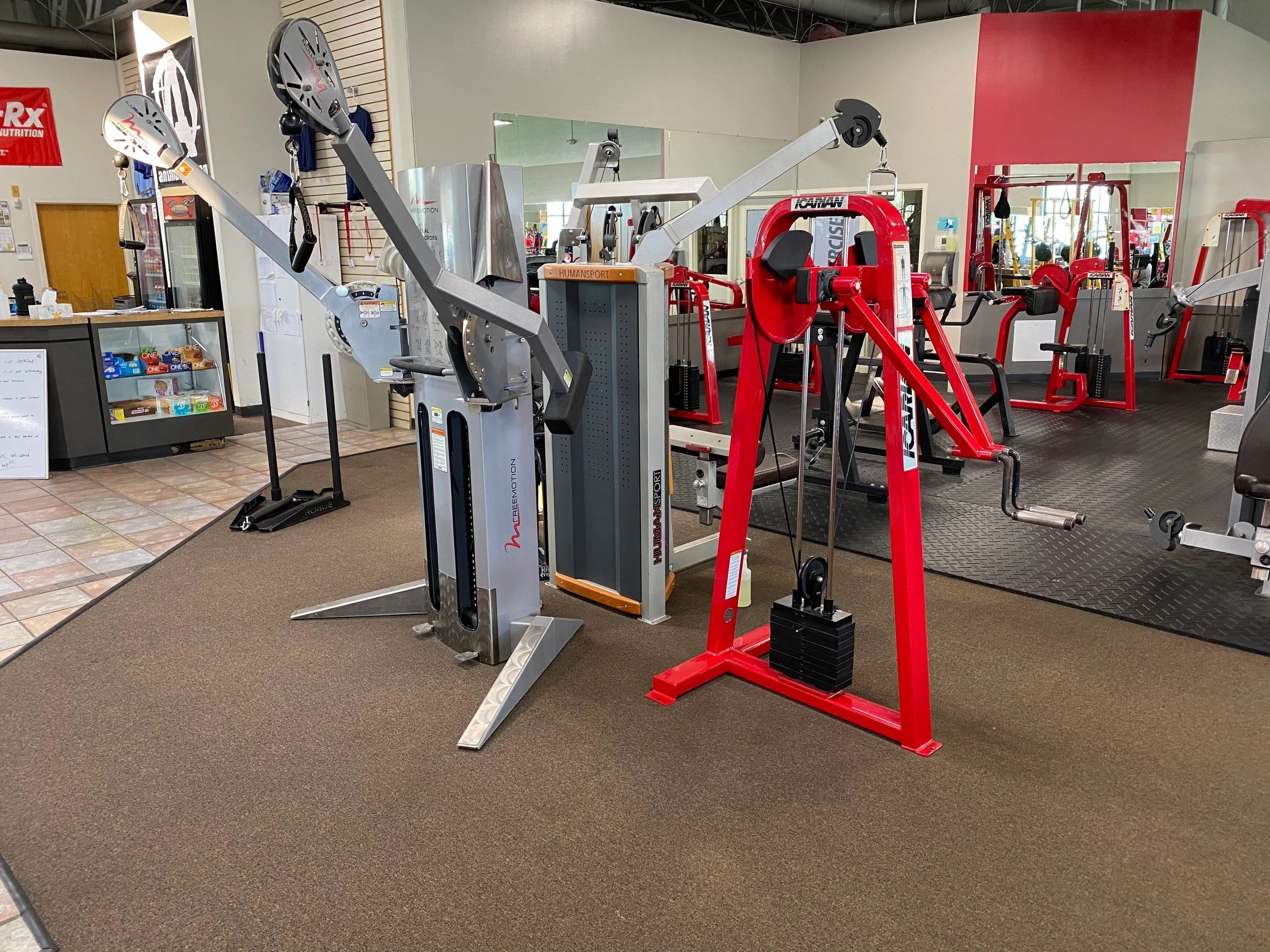 Gym equipment including a Precor exercise machine and a red cable machine on a carpeted floor. Mirrors and more equipment are visible in the background.