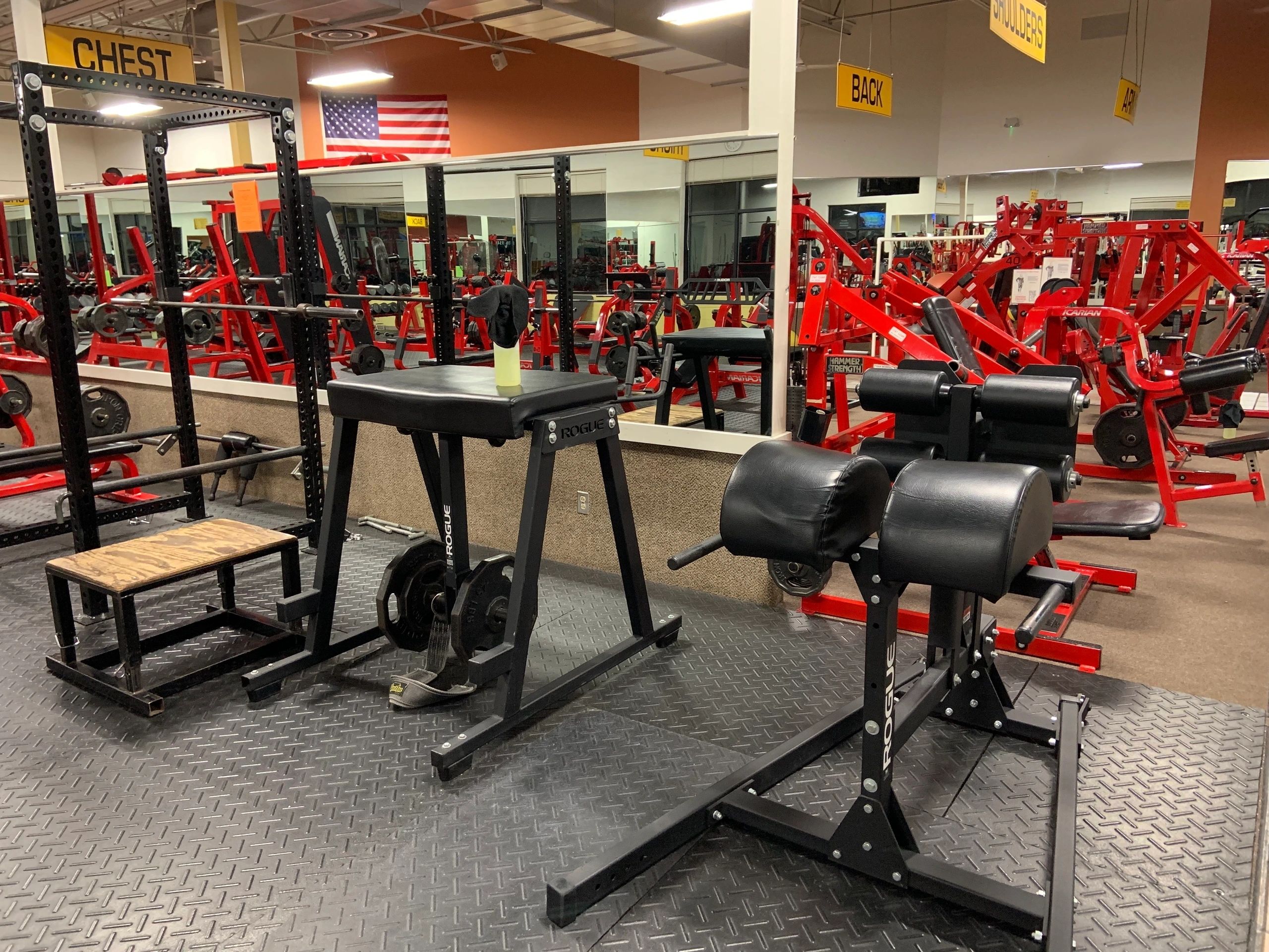 Gym with various exercise equipment, including weight benches, a squat rack, and mirrors on the wall.