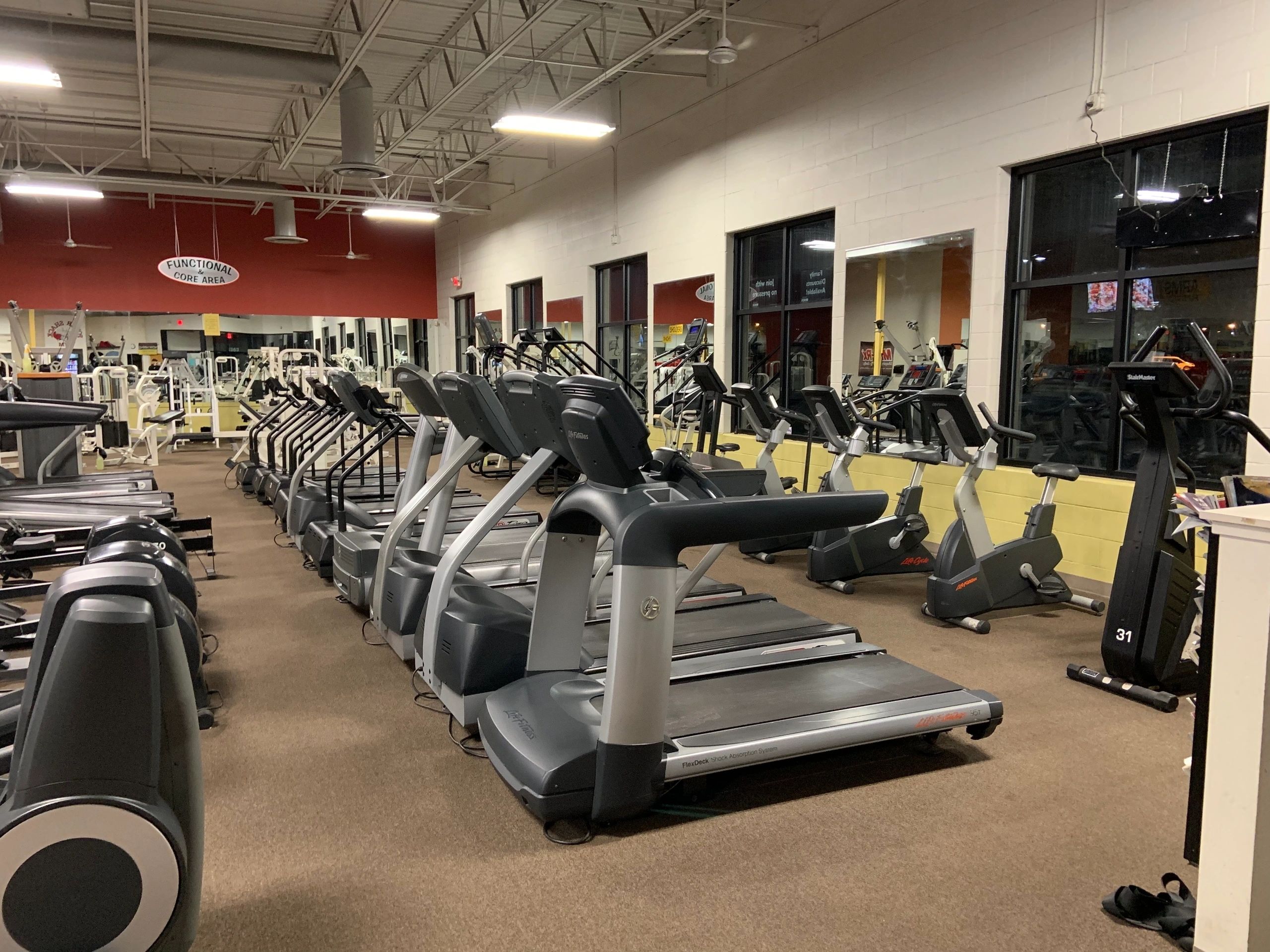 Rows of treadmills and exercise bikes lined up in a gym with red and white walls, large windows, and fluorescent lighting.