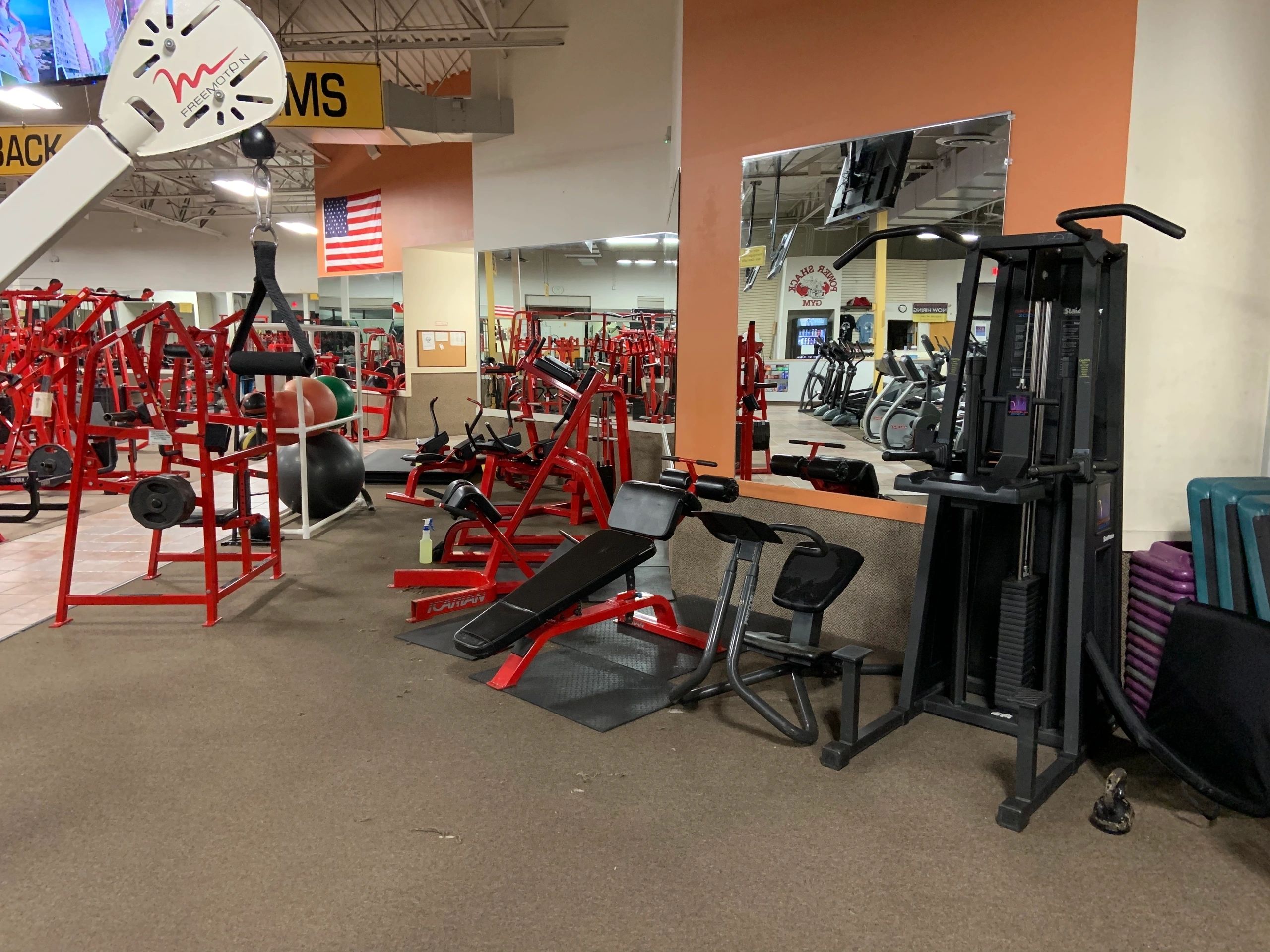A gym with red and black exercise equipment, including benches, a cable machine, and an American flag on the wall.