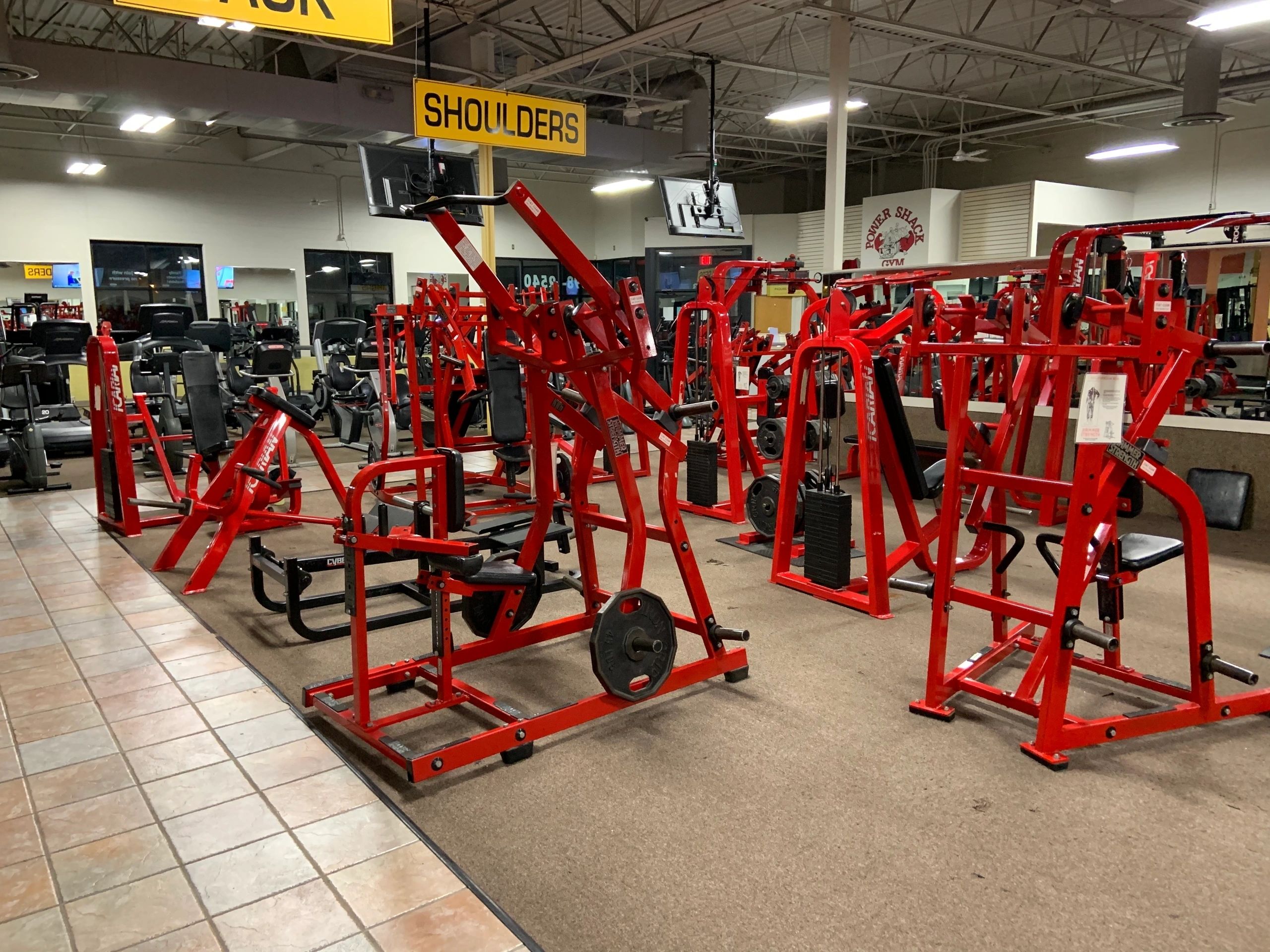 A variety of red exercise machines are arranged in a gym, with a sign overhead labeled "SHOULDERS.