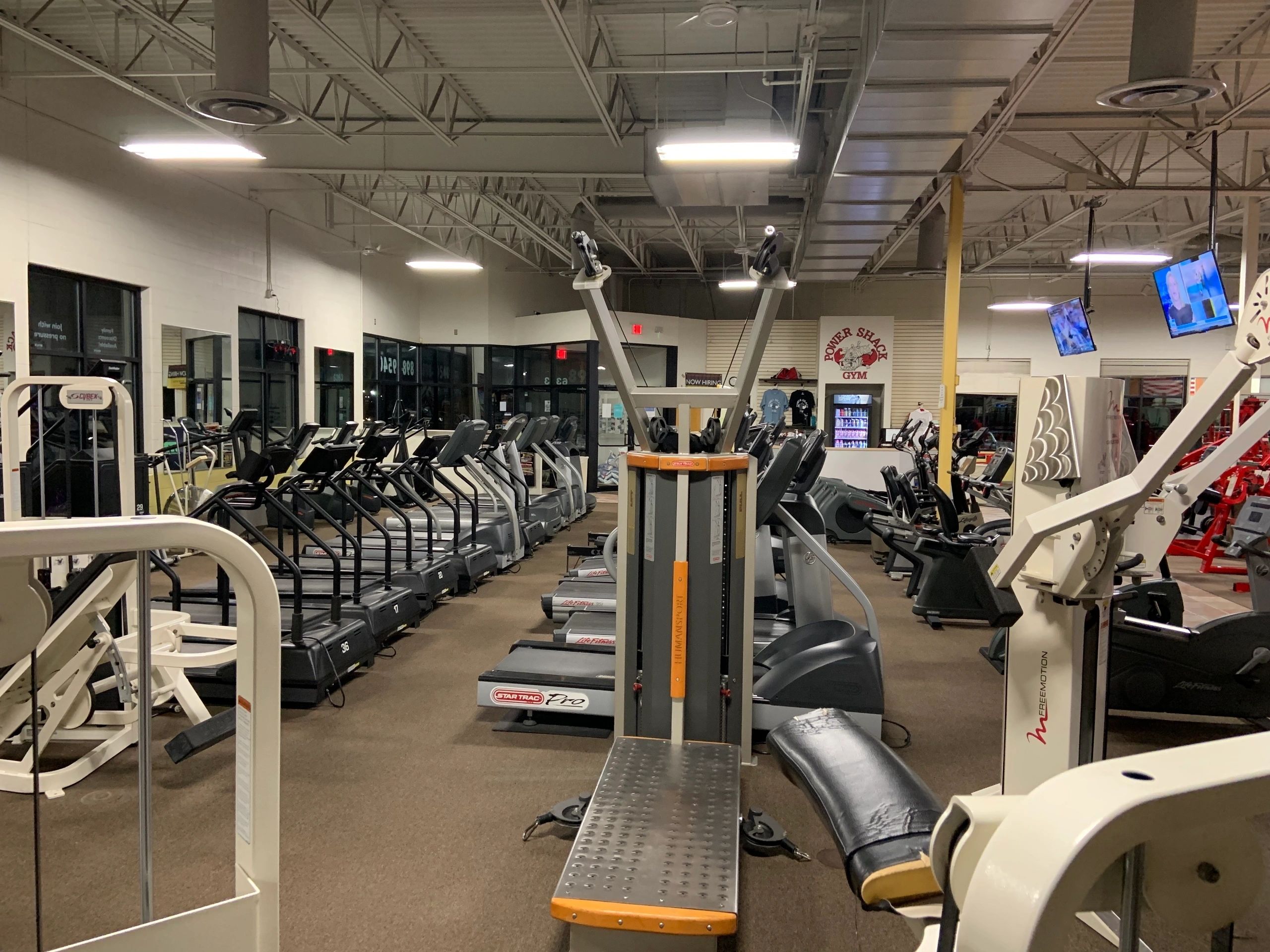 A gym interior with various exercise machines including treadmills, ellipticals, and strength equipment. The area is well-lit with overhead lights.