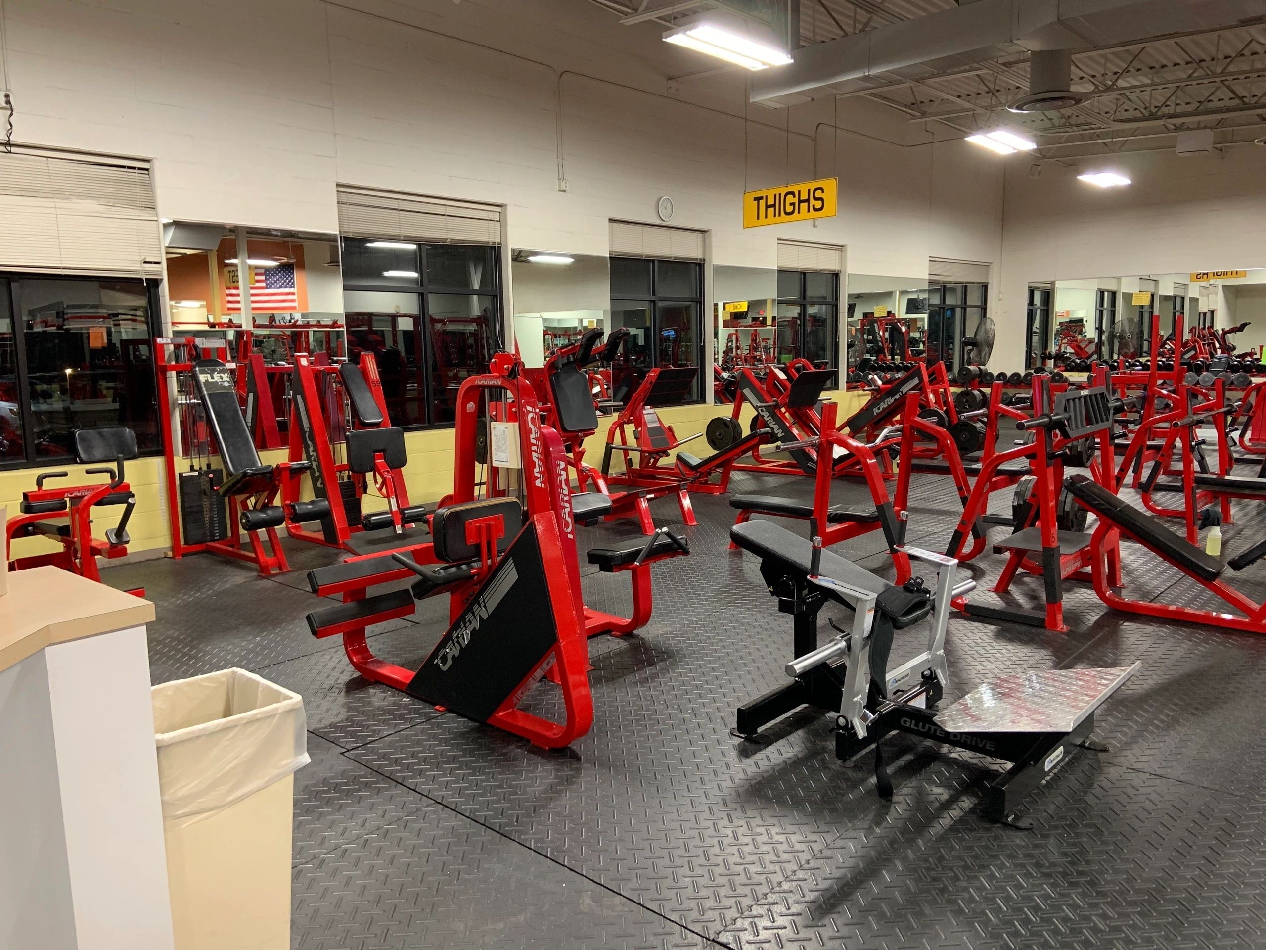 A gym interior with red and black exercise equipment, including weight machines and benches, on a black mat floor. A sign for "Thighs" is hanging from the ceiling.