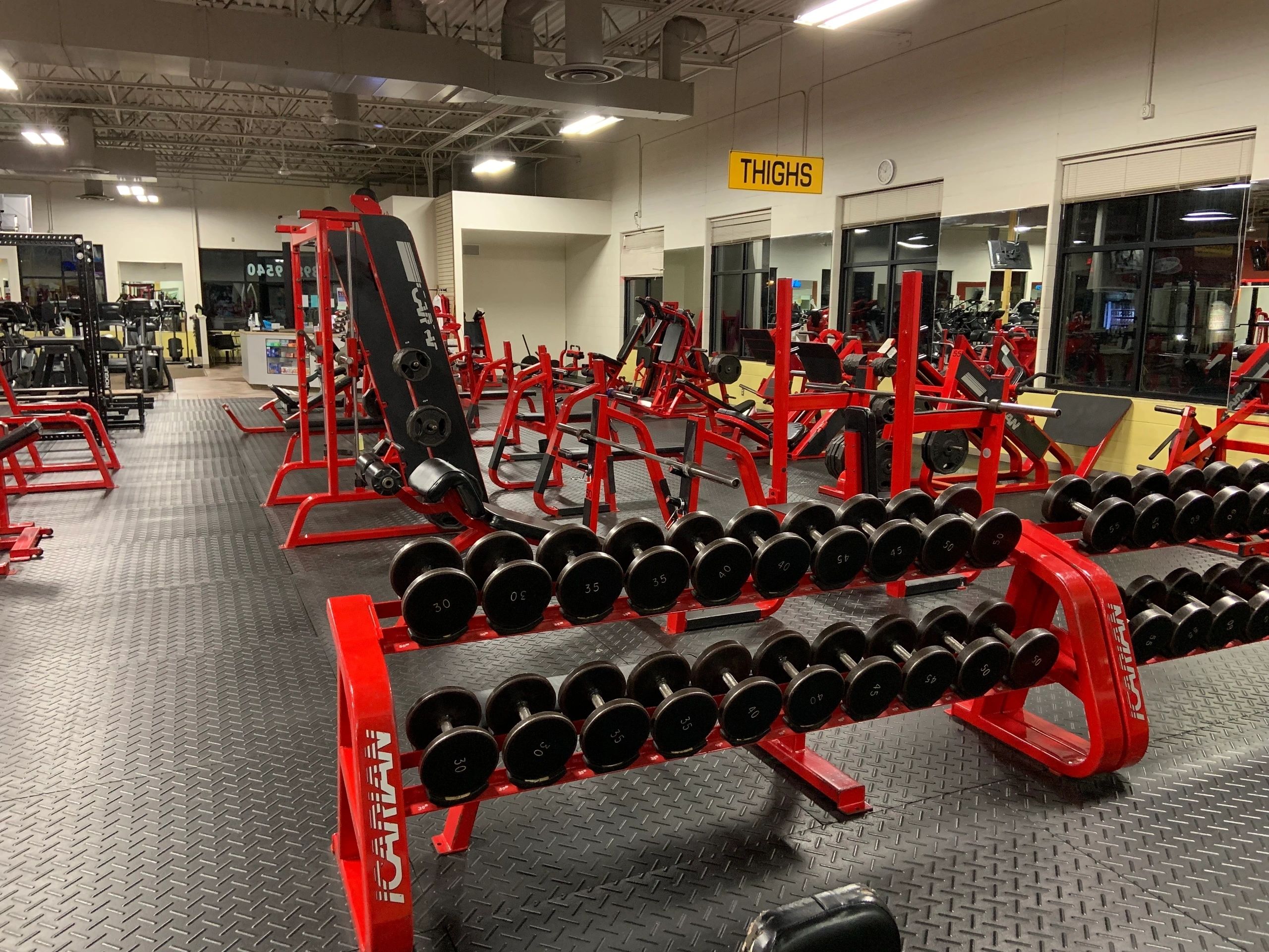 A gym interior with rows of red weightlifting machines, dumbbells, and a sign reading "THIGHS" on the wall.