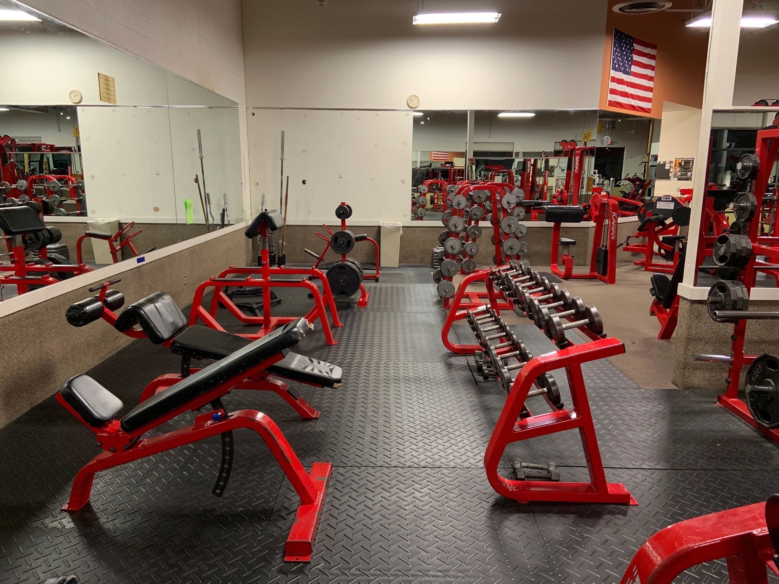 A gym with red equipment, including benches, dumbbells, and weights, on black flooring. An American flag hangs on the wall above mirrors.