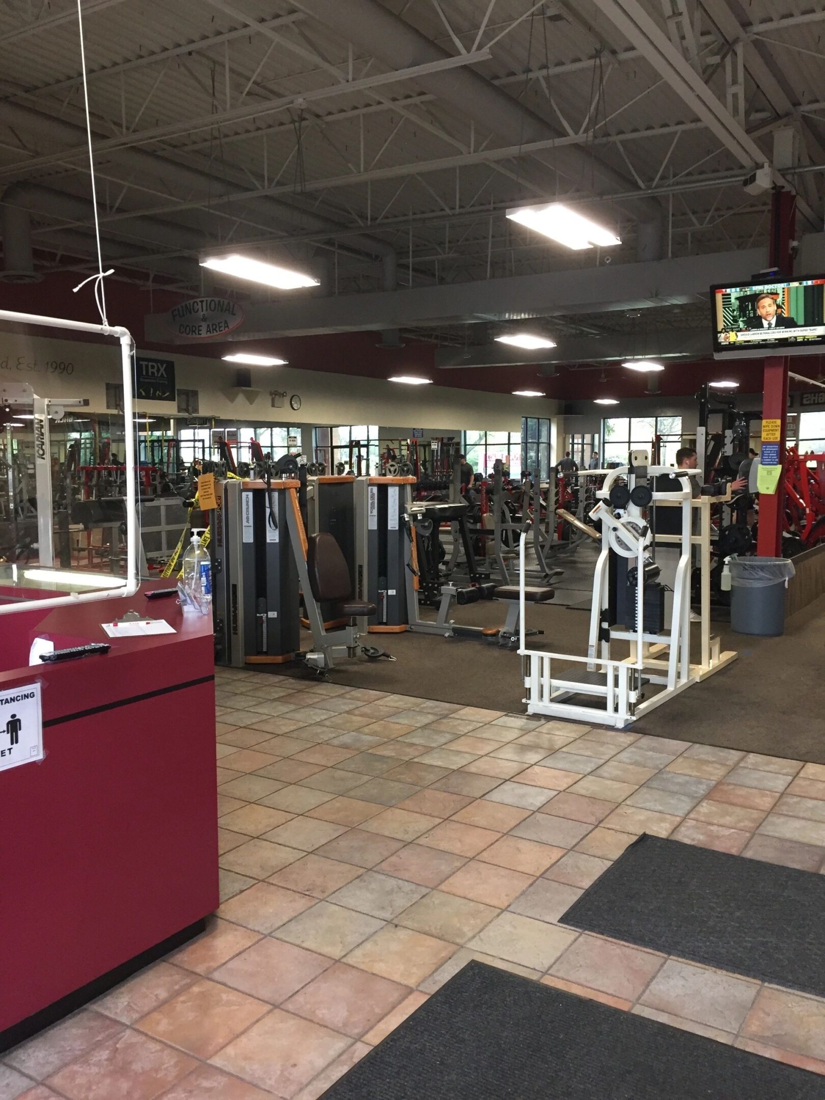 Empty gym with various exercise machines, a TV playing in the corner, and a red reception counter. Ceiling lights illuminate the spacious area.
