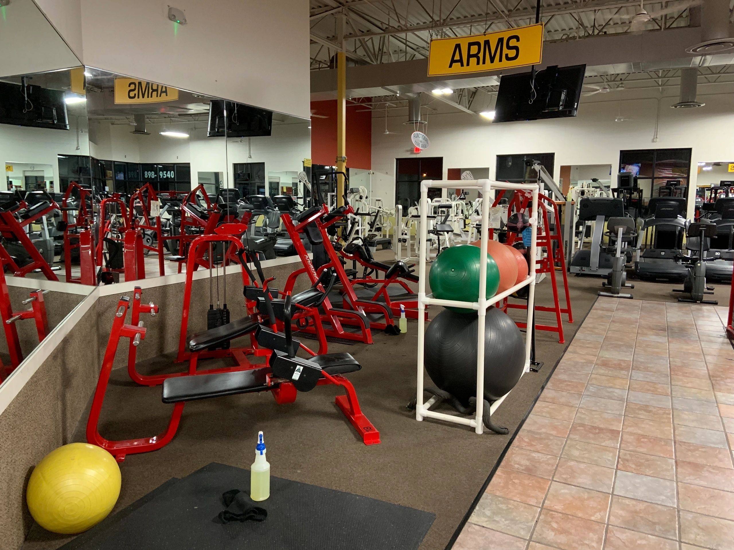 Gym interior with red exercise machines, resistance balls, and a mirror. A sign labeled "ARMS" hangs above. Cleaning spray and cloth are on a mat in the foreground.