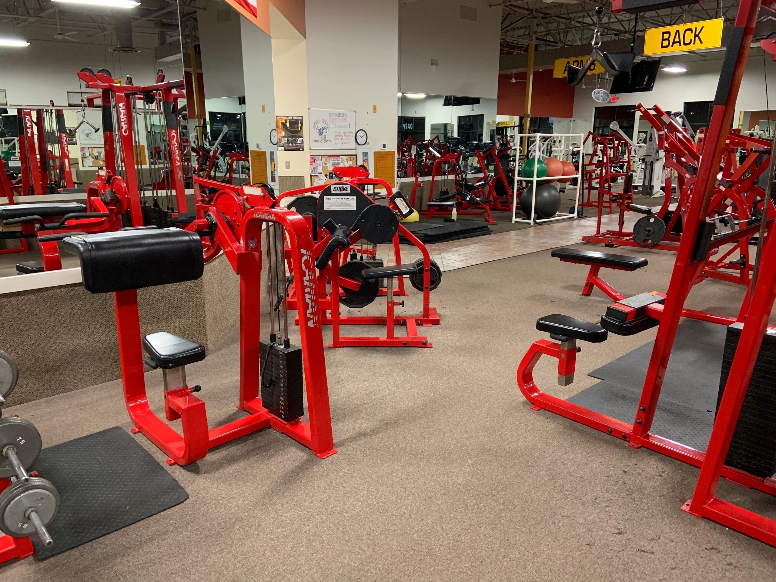 A gym with red weight machines, mirrors on the wall, and a rack of dumbbells on the left. The space is well-lit and tidy.