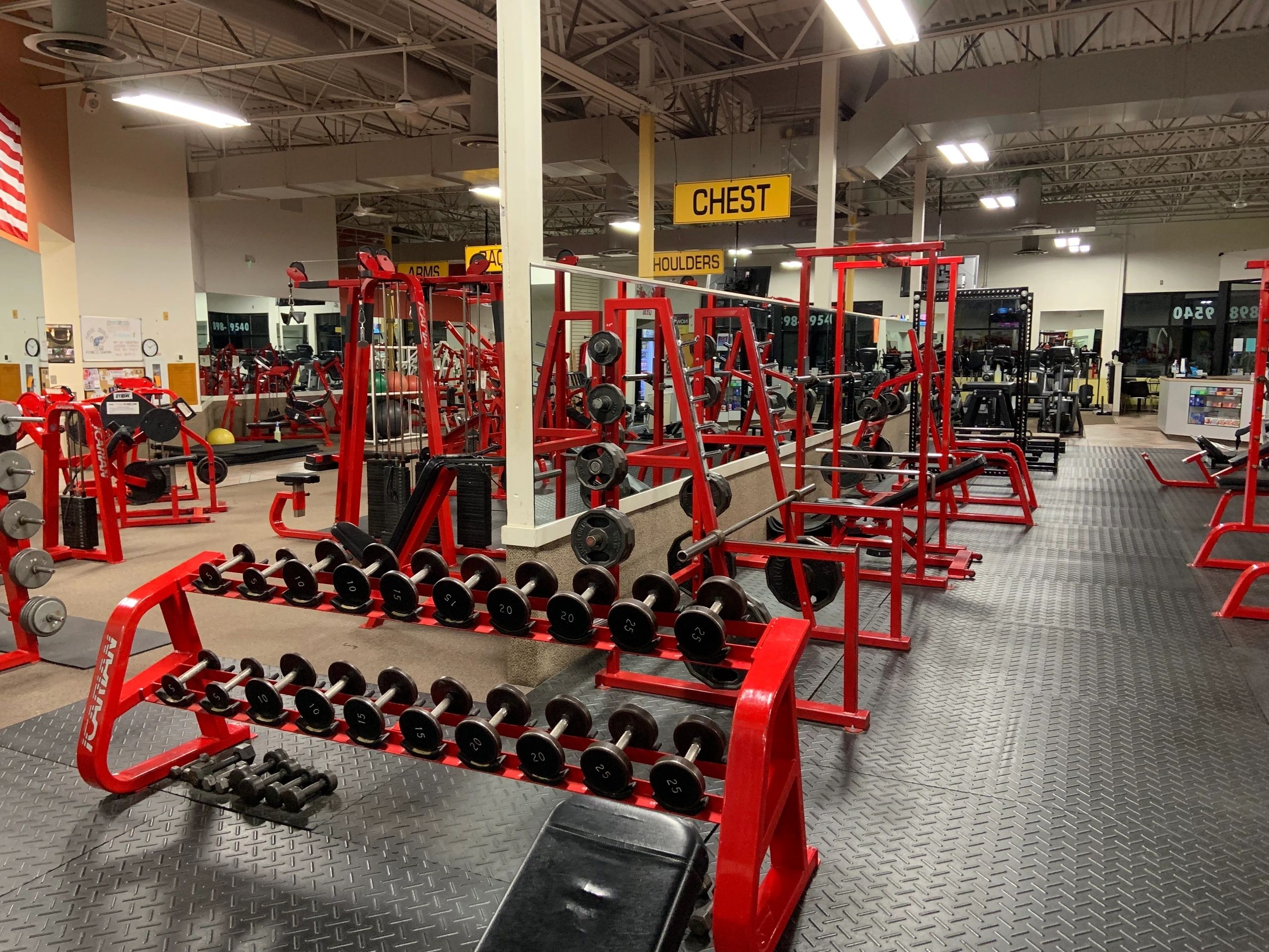 A gym with red weight racks, dumbbells, and various exercise equipment on a black rubber floor. A sign labeled "Chest" hangs from the ceiling.