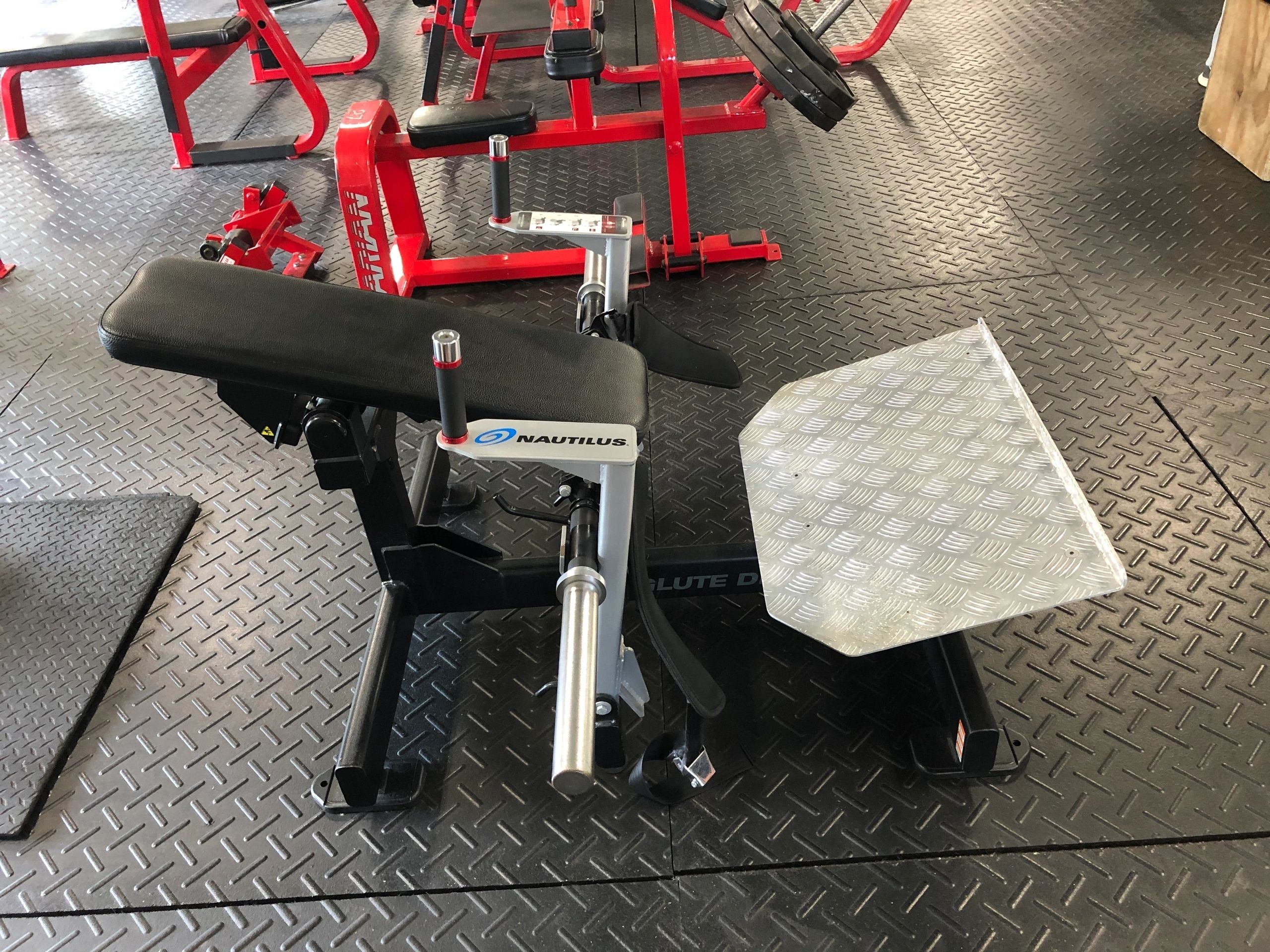A gym machine with black padding and a silver diamond-patterned footplate on a rubber mat floor. Red equipment is visible in the background.