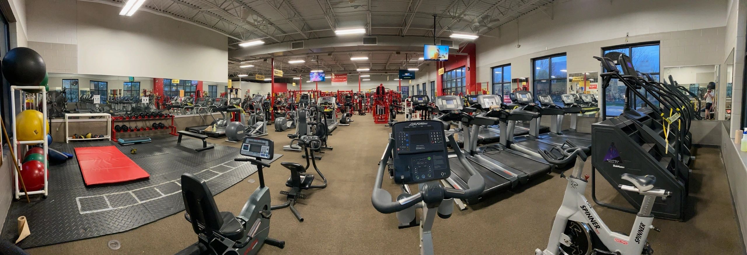 Panoramic view of a gym with cardio machines on the right, weightlifting equipment on the left, and exercise balls in the corner. Large mirrors cover the walls.