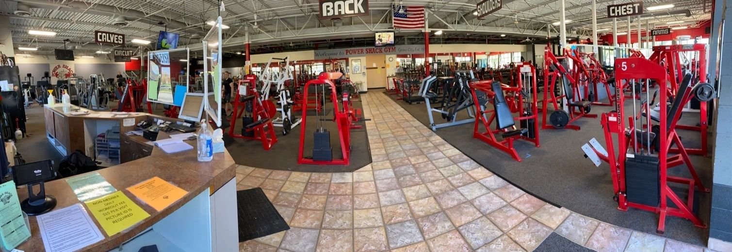 Wide-angle view of a gym with various weightlifting machines, a counter with papers and sanitizer, and signs indicating workout areas. An American flag is hanging from the ceiling.