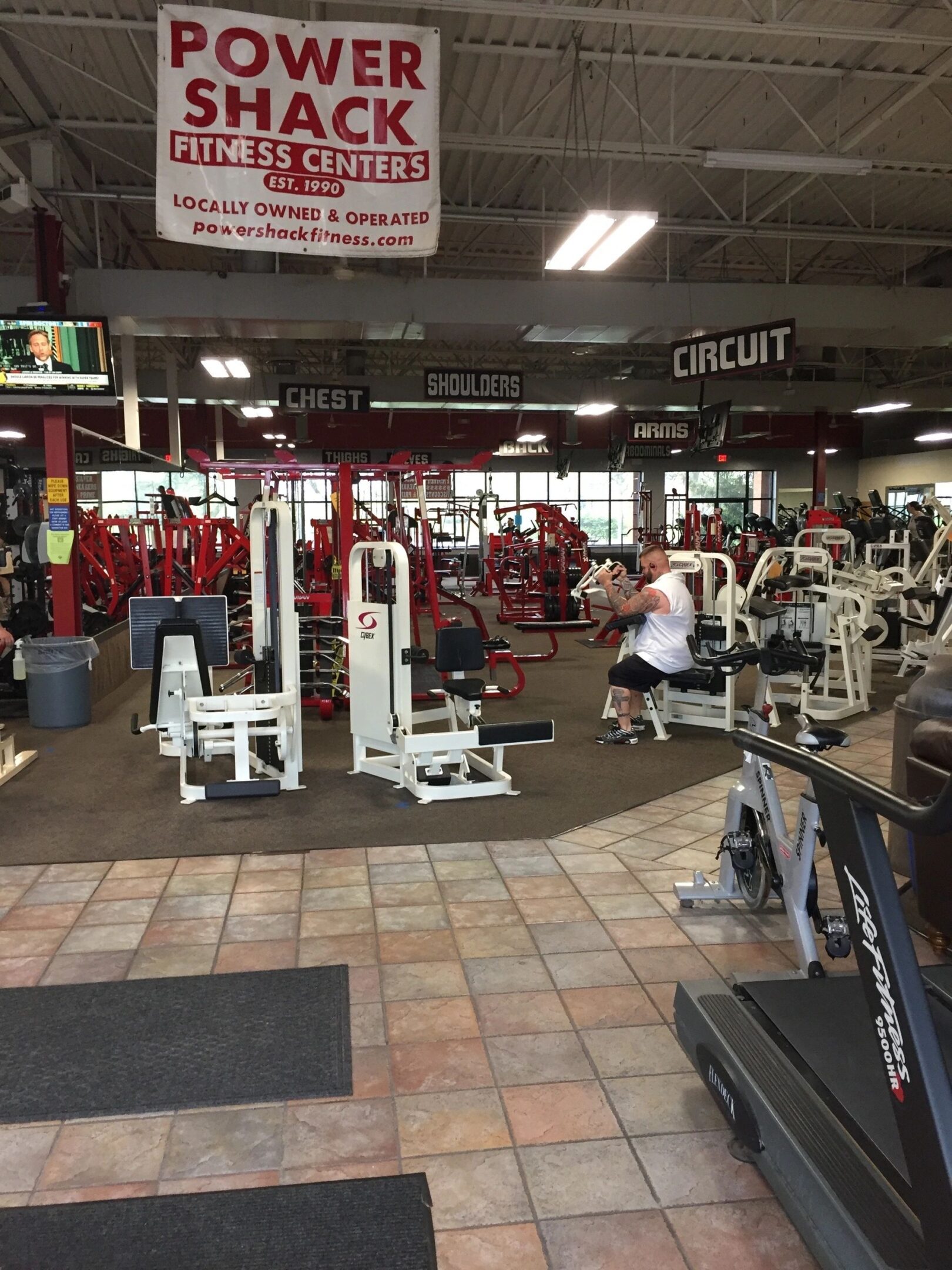 A gym interior with various exercise machines and equipment. A person is sitting on a machine. Signs for different workout areas like "Chest" and "Shoulders" are visible.