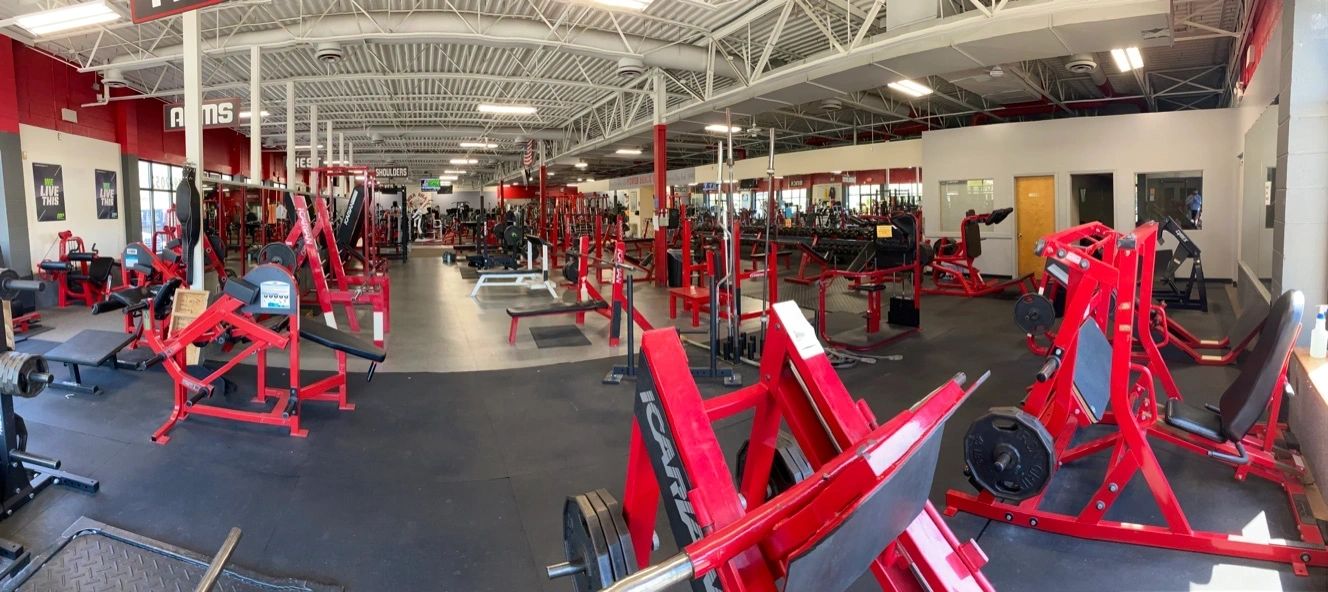 Gym interior with various red and black workout equipment, including benches, machines, and weights, under a ceiling with visible pipes and lights. Walls are red and white.