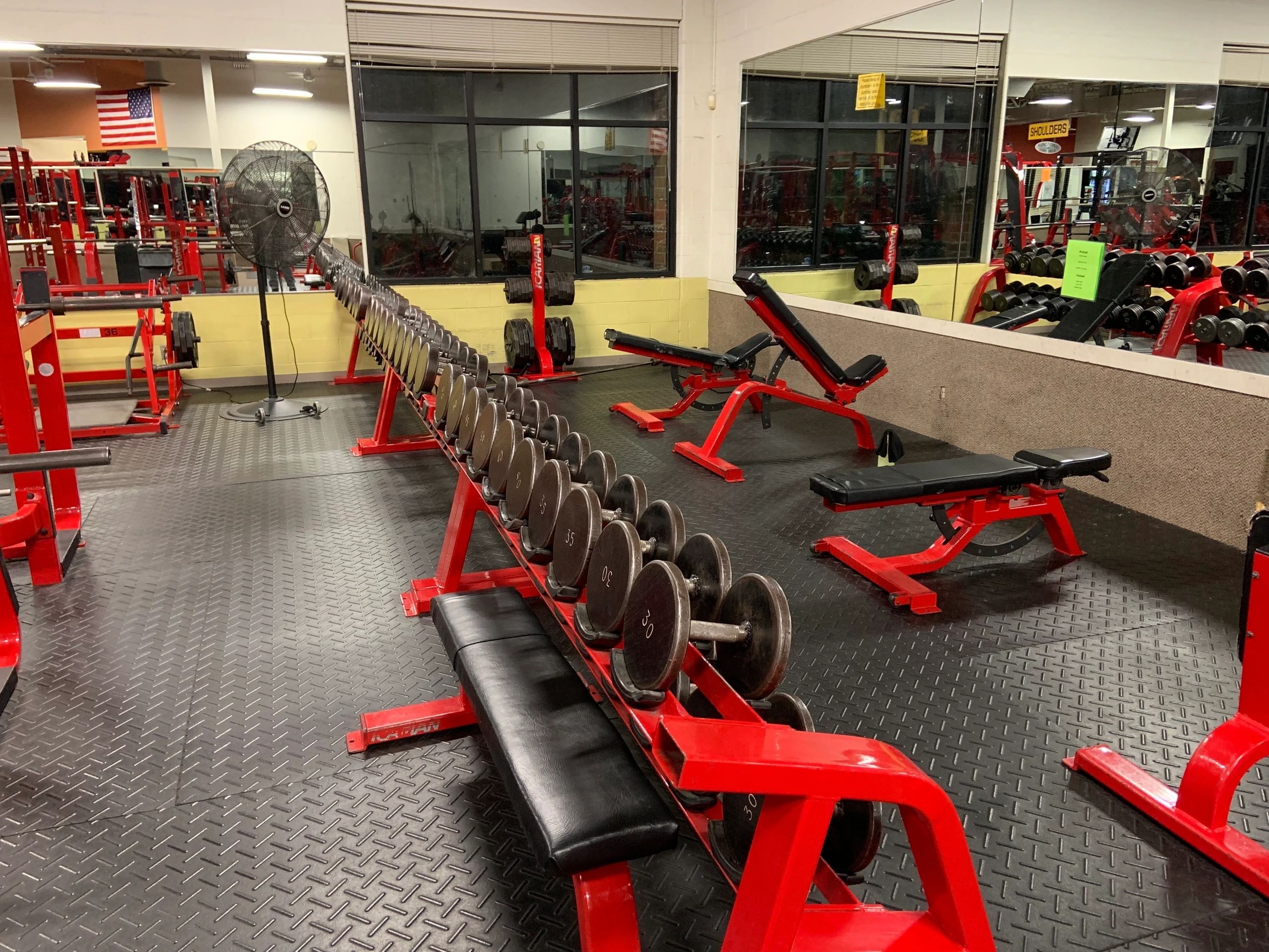 A gym with rows of dumbbells on red racks, weight benches, and mirrored walls. A large fan is visible in the background.