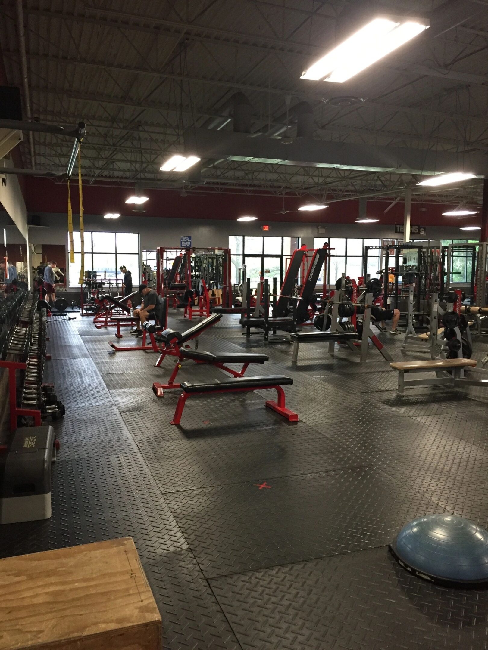 Spacious gym interior with various exercise equipment, including benches, weights, and machines. Several people are working out. Large windows on one side.