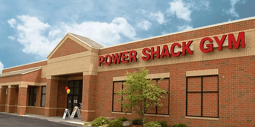 Brick building exterior with "POWER SHACK GYM" in red letters, clear sky overhead, small tree and balloons near the entrance.