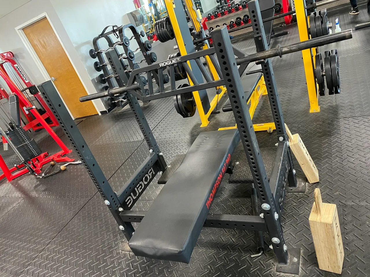 A gym bench press setup with a barbell, weight plates, and wooden blocks on a rubber mat floor. Red and yellow gym equipment is visible in the background.